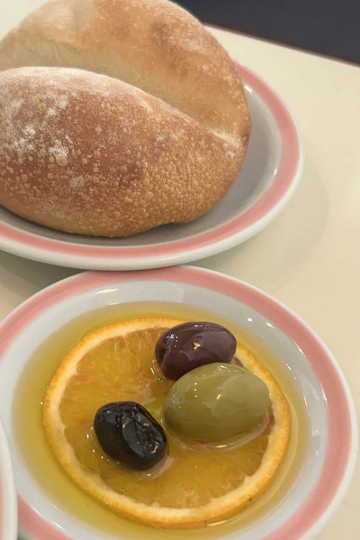 Bread and oil El Preferido serves. A slice of orange in a plate of olive oil. Green, black and plum color olives are on top of the orange slice.