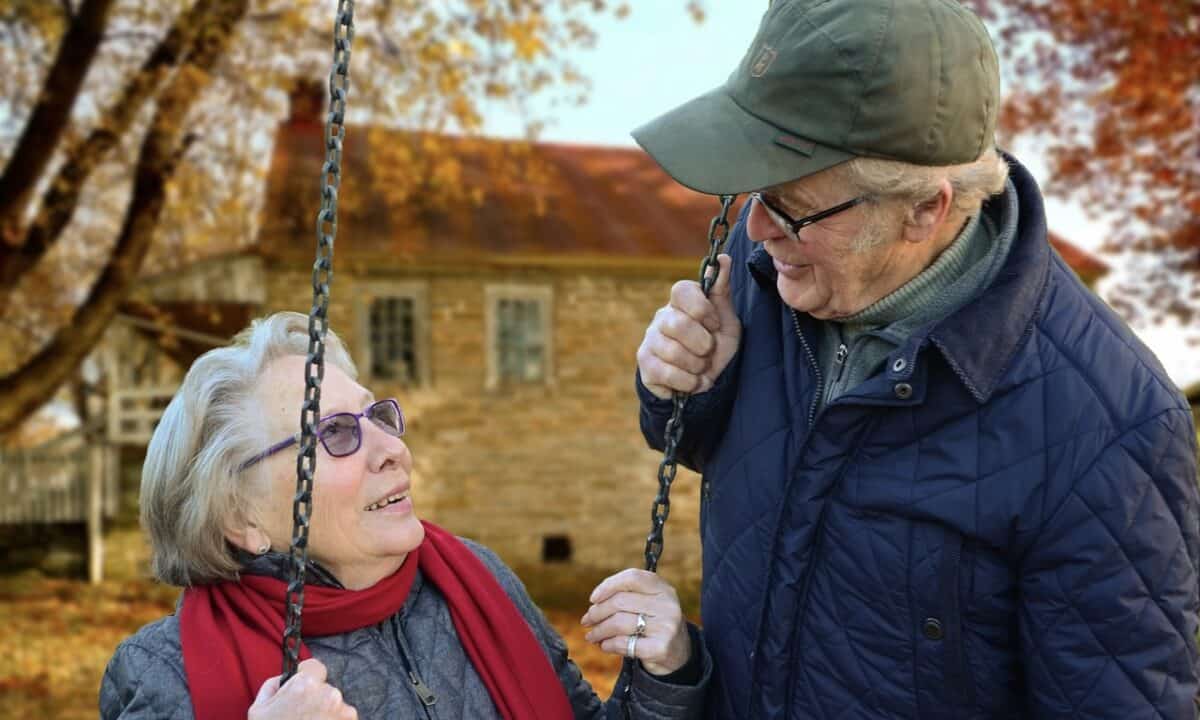 Old lady with red scarf sitting on a swing while her husband looking at her. Both are smiling.