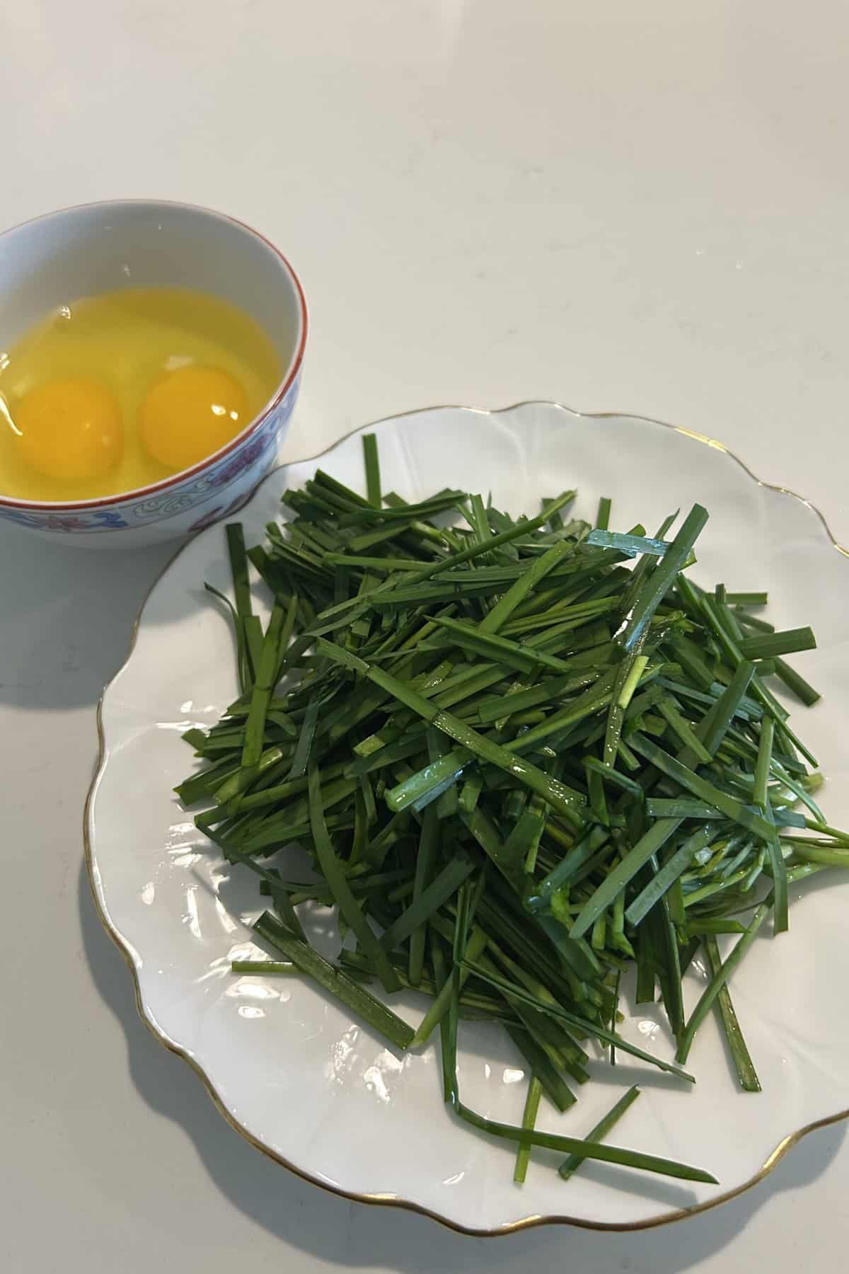 Two eggs in a bowl and Chinese chives cut up on a white plate ready to be cooked